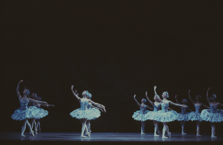 Ballet dancers performing on stage in silver, shimmering tutus during a live production of the Cinderella ballet against a dark backdrop.